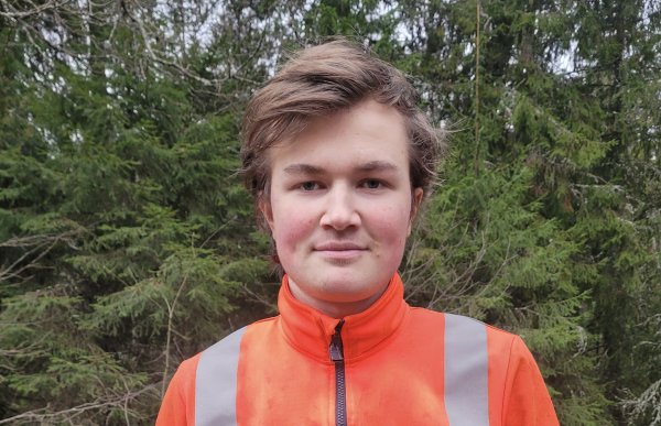 En ung mann med brunt hår smiler. Bak han er det skog. Han har på seg en oransj genser med refleksstriper. 