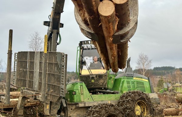 En stor grønn hogstmaskin holder oppe flere tømmerstokker. I hogstmaskin sitter det en mann med svart genser og brunt hår. Hogstmaskinen har kjettinger på hjulene og det er overskyet himmel i bakgrunn.