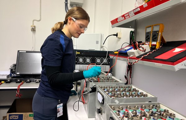 Dame i et verksted jobber med noen store metallbokser på en benk, mens hun bruker vernebriller og hansker for å være trygg