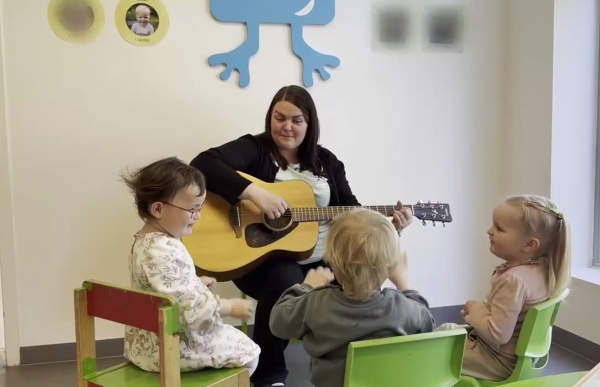 En kvinne med brunt, halvangt hår sitter på en stol og spiller gitar for tre små barn som sitter rundt henne på lave stoler. Hun har på seg en lys grønn t-skjorte og en svart cardigan. Barna ser på henne og deltar i aktiviteten. På veggen bak henger det bilder og dekorasjoner.