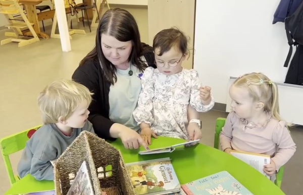 En kvinne med halvlangt, brunt hår sitter ved et lite bord sammen med tre små barn og ser på en bok  sammen med dem. Hun har på seg en lys grønn t-skjorte og en svart cardigan. Barna sitter tett rundt bordet, og det ligger flere barnebøker foran dem.