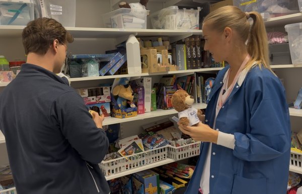 En kvinne i blå frakk står sammen med en annen person foran hyller fylt med leker, bøker, spill og bamser. Kvinnen holder en liten teddybjørn i hendene mens de ser på innholdet i hyllene.