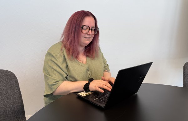 En kvinne med rødt hår og store briller sitter ved et rundt bord og arbeider på en bærbar PC. Hun har på seg en lys grønn topp og ser ned på tastaturet mens hun skriver. Bildet er tatt i et enkelt møterom med lyse vegger og mørke stoler.