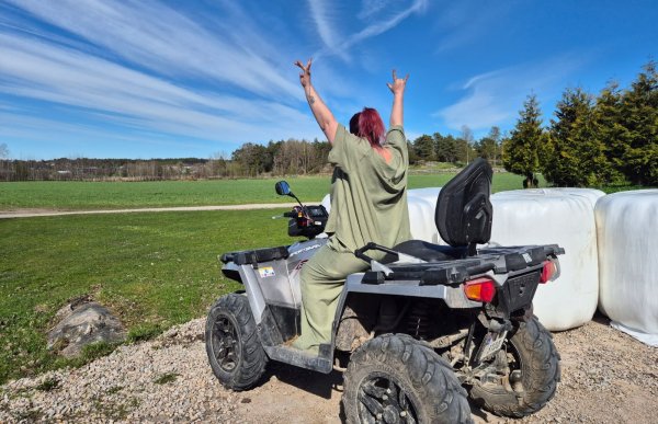 En kvinne med rødt hår sitter på en firhjuling ute på et jorde en solrik dag. Hun har ryggen mot kamera og løfter armene i været. Rundt henne er det grønne jorder, trær og store hvite rundballer. Himmelen er blå med lange, lyse skyer.