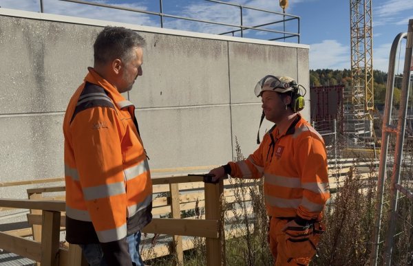 To menn i oransje arbeidsjakker med refleks står og snakker sammen på en byggeplass. Den ene har kort, grått hår, mens den andre har vernehjelm med hørselsvern. I bakgrunnen er det en betongvegg, stillaser og en byggekran.