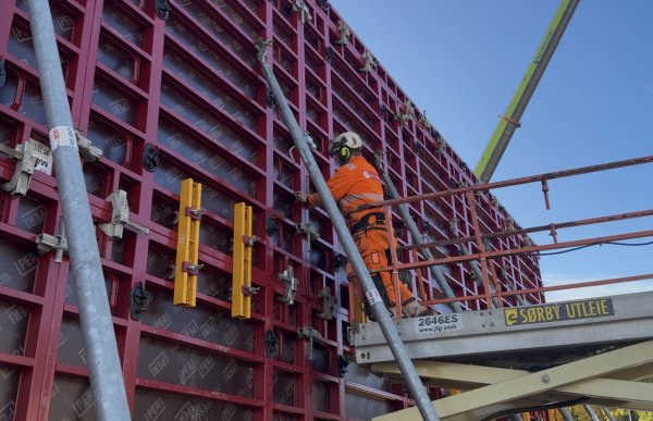 En mann i oransje arbeidsklær, vernehjelm og sikkerhetssele står på en arbeidsplattform høyt oppe på en byggeplass. Han arbeider med å  justere deler av en stor, rød forskalingsvegg. Flere skrå metallstøtter holder konstruksjonen på plass, og en kran er synlig i bakgrunnen.