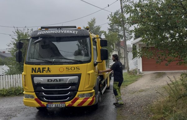 En mann står ved siden av en stor gul bergingsbil fra Redningsverket på en våt vei. Han har på seg mørk T-skjorte og arbeidsbukse med gule felt og refleks. Rundt bilen er det bolighus, trær og hager.