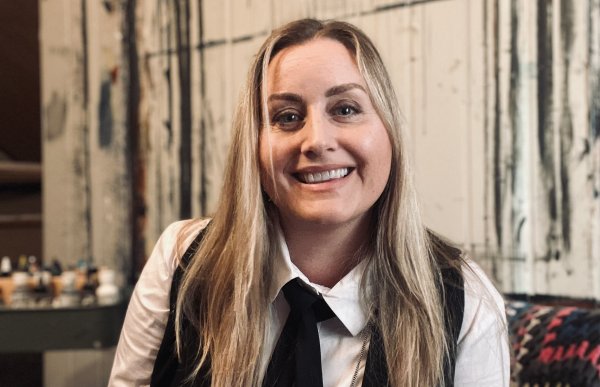 En dame med langt blondt hår smiler. Hun har på seg hvit skjorte, med svart vest og svart slips. Hun sitter på en brun stol og det ligger et lappeteppe bak hun. I bakgrunnen er det maling og en vegg som har blitt malt på.