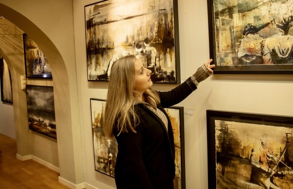 En dame med langt, lyst hår og svart åpen genser med ullgenser under står sidelengs å peker på et malleri på veggen. Det er flere mallerier på veggen bortover. Malleriene ligner på hverandre og de har alle sorte rammer.