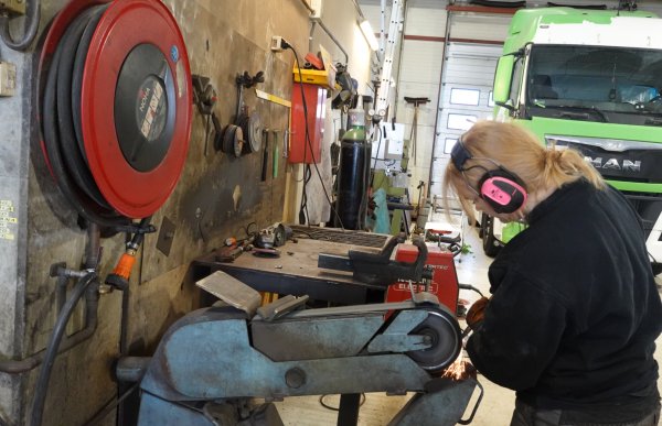 Jente med blond hestehale, rosa hørselvern og svart genser sliper et verktøy i en slipemaskin.