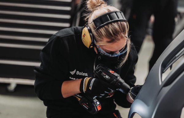 Dame med hørselvern, vernebriller, hansker og svart genser står med et borr mot en bil