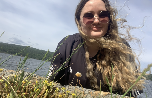 En kvinne tar en selfie utendørs ved vannet. Hun ligger eller lener seg lavt mot bakken, med kameraet plassert nær ansiktet. Hun har langt, bølgete hår som blåser i vinden, og hun bruker runde solbriller med mørke glass og en svart treningsgenser med hvite striper på ermene. Foran henne i forgrunnen er det gress og små gule blomster, og bak henne ligger en innsjø med skogkledde åser i bakgrunnen. Himmelen er lys, og det ser ut som en solrik og litt vindfull dag.