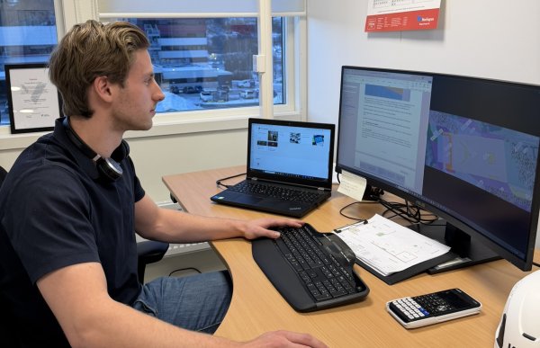 Roy Martin Adolfsen Stiberg Pettersen sitter på kontoret og jobber med en tegning på pc-en og noen andre programmer. Det er en stor pc-skjerm, tastatur og og en liten, bærbar pc ved siden av. Han har på seg svart t-skjorte og på veggen henger det en kalender. 