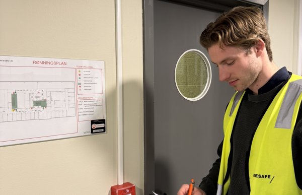 En mann med gul refleksvest over en svart genser står foran en dør med rundt glass. Over døra henger det et nødutgangskilt, og på veggen henger det en brannarlam og en rømningsplan.