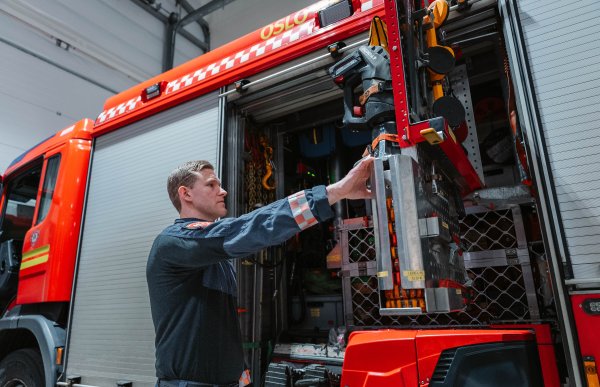 En mann i brannuniform står ved siden av en brannbil med åpen dør. Han strekker hånden opp mot innsiden av døren, hvor det henger utstyr og verktøy.