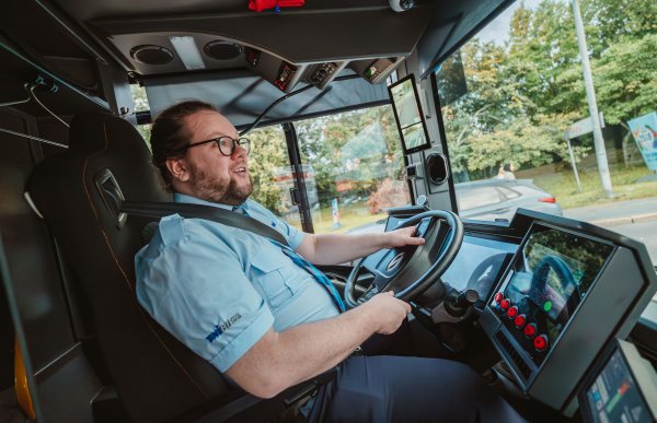 En mann med langt hår og skjegg, iført uniform med lys skjorte og mørk bukse, sitter i førersetet på en buss. Bussen er i bevegelse, og han ser konsentrert ut mens han kjører.