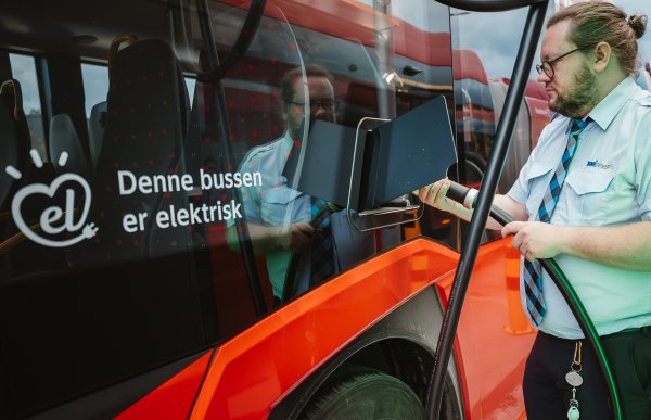En bussjåfør i uniform plugger laderen inn i en elektrisk buss. Bussen er rød, og på siden står det "denne bussen er elektrisk".
