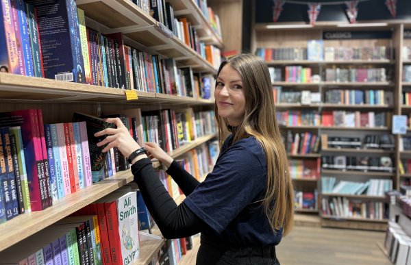 Brunhåret dame med blå genser står foran en bokhylle og skal til å ta ut en bok. Det er bokhyller overalt i rommet.