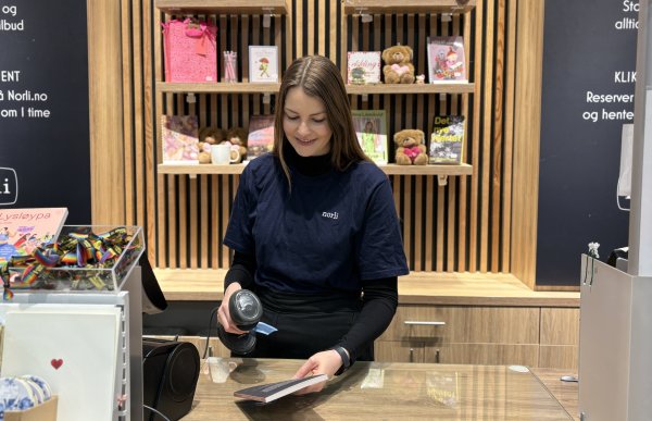 Brunhåret dame med blå genser står bak en kasse og scanner en vare. 