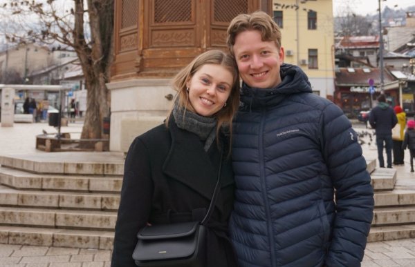 Kvinne med langt blondt hår, iført en lang svart kåpe, står sammen med en gutt med kort blondt hår og mørkeblå jakke. De poserer under en tur i Sarajevo.