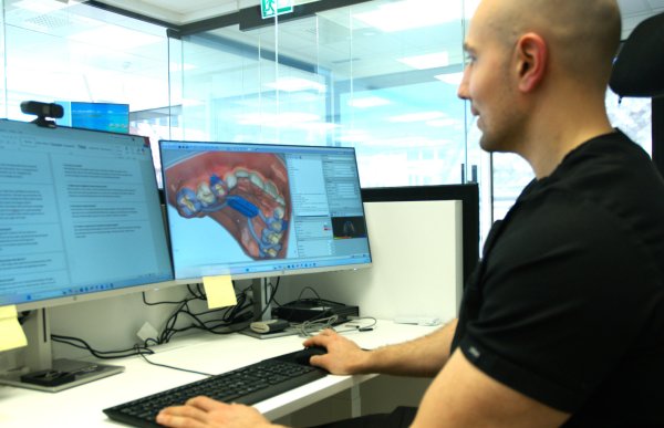 Kjeveortopeden i svart uniform sitter foran to dataskjermer og justerer reguleringen vi ser på den ene skjermen.
