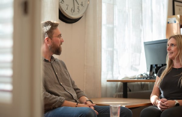 To personer sitter i samtale rundt et bord på et kontor. En mann med skjegg og en kvinne med langt lyst hår ser på hverandre og smiler. I bakgrunnen er det et vindu, en klokke og en dataskjerm.