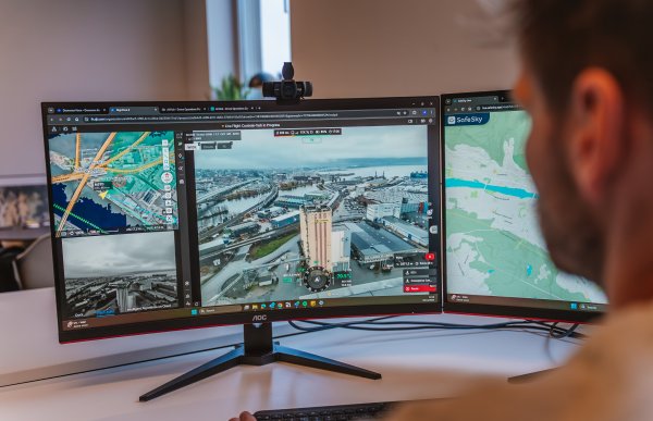 En skjerm på en kontorpult viser et kartutsnitt, et onboard-kamera fra en drone og et dronekamera som viser bygninger sett fra luften. I forgrunnen sitter en mann og ser på skjermen.