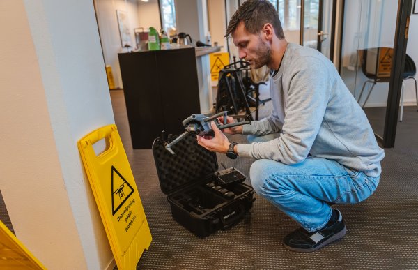 En mann med skjegg, iført olabukse og en lysegrå genser, sitter på huk og setter sammen en liten drone. Ved siden står et gult skilt med teksten "droneflygng pågår".