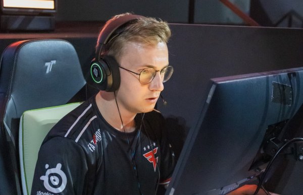 En e-sportutøver sitter i en gamingstol foran en dataskjerm i en turneringsarena. Han har på seg headset med mikrofon og runde briller. Han ser konsentrert på skjermen. Han har på seg en svart drakt med laglogo og sponsormerker.