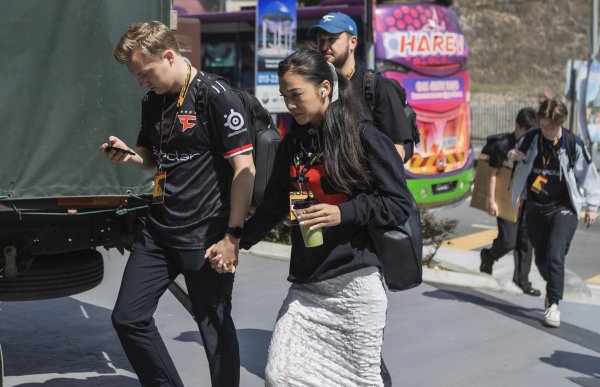 Flere personer i e-sportdrakter går ute i sollys på vei til eller fra et arrangement. En mann og en kvinne går hånd i hånd i forgrunnen, begge med akkrediteringskort rundt halsen og utstyr på ryggen. I bakgrunnen står en fargerik buss og flere andre deltakere går samme vei.