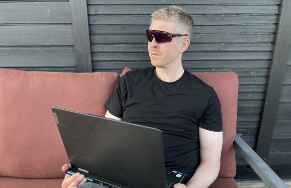 En mann med svart t-skjorte, grå shorts og svarte solbriller sitter på en rød utesofa og jobber med en svart PC på fanget. Bakgrunnen er en svart husvegg.