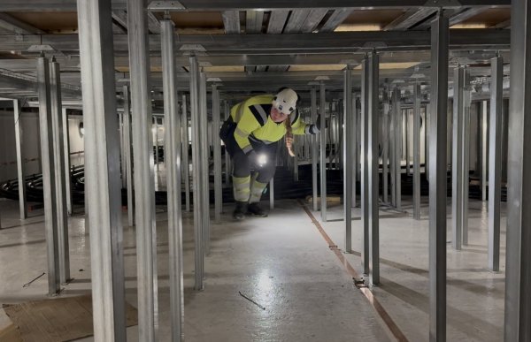 En kvinne i gul arbeidsgenser med refleks, mørk arbeidsbukse og hvit vernehjelm beveger seg fremover i en lav konstruksjon med mange metallstøtter. Hun går fremoverbøyd og lyser foran seg med en lommelykt.
