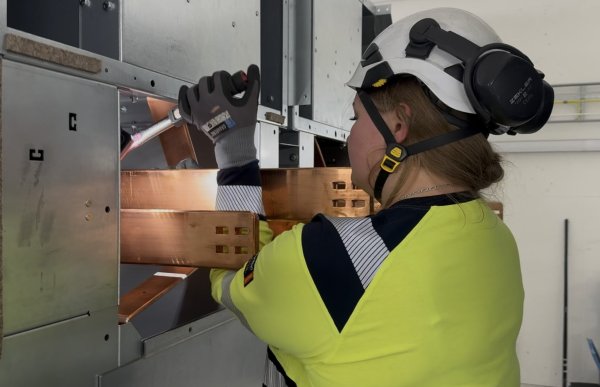 En kvinne i gul arbeidsgenser med refleks, hvit vernehjelm med hørselsvern og arbeidshansker arbeider inne i et elektrisk anlegg. Hun holder et verktøy mot en kobberfarget metalldel inne i et åpent elektroskap.