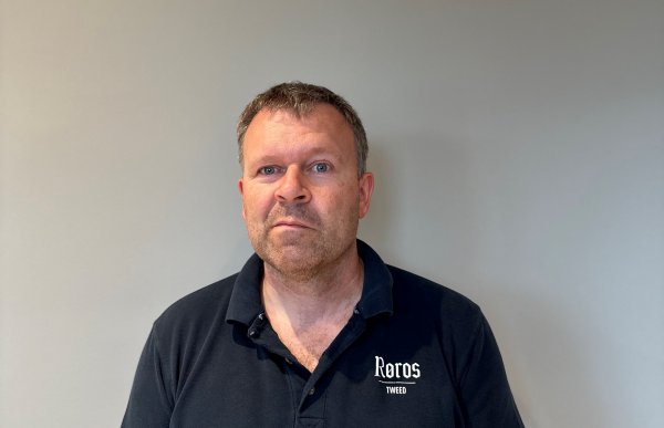 Mann med svart t-skjorte står inntil en grå vegg og ser inn i kameraet.