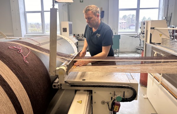 Mann i svart t-skjorte står ved en vev. Garnet er lyst og brunt. Det er to vinduer i rommet.