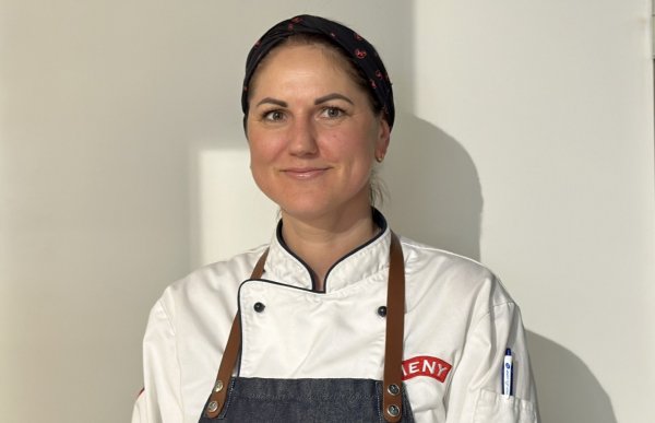 En kvinne med hvite arbeidsklær og blått forkle. På arbeidsklærne står Meny-logoen. Kvinnen har oppsatt hår og et svart tørkle på hodet. Hun smiler mot kameraet.