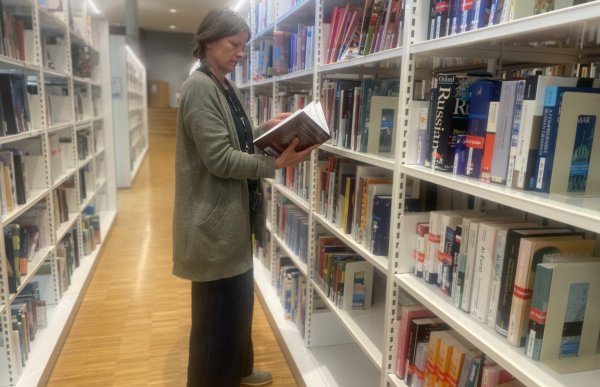 Dame med grå cardigan står mellom to reoler på et bibliotek og leser i en av bøkene.