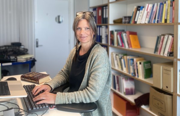 Dame med grå cardigan og briller på hodet sitter foran en pult med to pc-er og skriver. I bakgrunnen er det en vegg med bokhyller og en hvit dør som leder ut av kontoret.