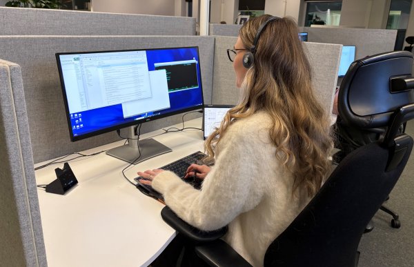 Dame med lyst hår og briller og headset sitter foran en pc-skjerm i åpent kontorlandskap