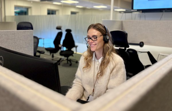 Dame med lyst hår og hvit genser sitter med headset, briller og ser på en pc-skjerm hun har foran seg. Det er et åpent kontorlandskap.