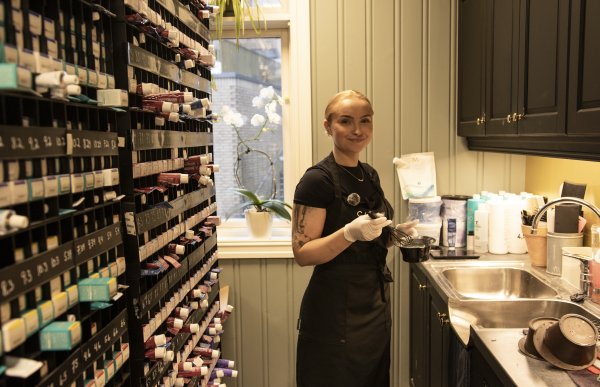 Frisøren står ved en arbeidsbenk med vask og skap rundt seg, og holder en bolle og en pensel til hårfarge. Personen har på seg mørkt forkle og engangshansker. På veggen ved siden av er det hyller fylt med mange hårfargetuber.