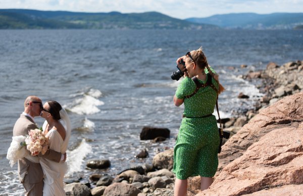 En blond kvinnelig fotograf i grønn mønstret kjole står på en svaberg i solen og tar bilde med kamera ned mot et brudepar som står i vannkanten. Bruden har på hvit brudekjole og holder en stor lys brudebukett, mannen har en beige dress. Foto