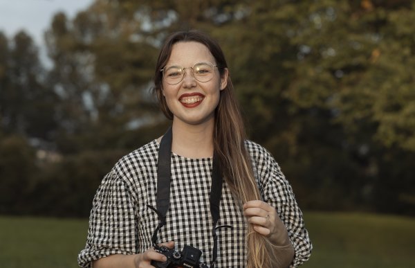 En kvinne med briller, mørkeblondt hår og rutete kjole. Hun har et kamera rundt halsen. Kvinnen smiler.