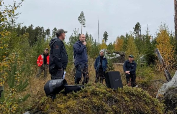 Fem menn står og sitter samlet ute i et skogsområde. En av mennene står i midten og snakker i en mikrofon, mens de andre lytter. Rundt dem vokser unge trær og annen vegetasjon, og på bakken står høyttalere og annet utstyr.