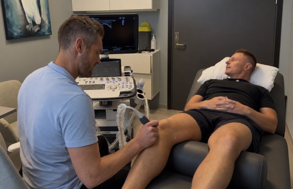 En mann ligger på en behandlingsbenk mens en annen mann undersøker kneet hans med ultralyd. Mannen som utfører undersøkelsen har på seg en lys blå T-skjorte, mens mannen på benken har svart T-skjorte og shorts. Ved siden av behandlingsbenken står et ultralydapparat med bilde av undersøkelsen på skjermen.