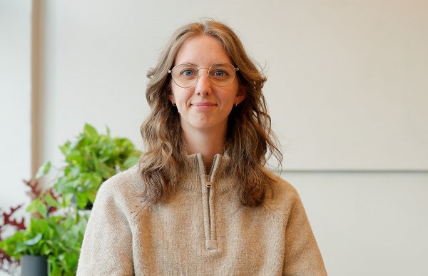 En kvinne med langt brunt hår og briller sitter i en stol og smiler til kamera. Hun har på seg ullgenser og jeans, og har hendene foldet i fanget. I bakgrunnen står en fargerik plante på et bord.