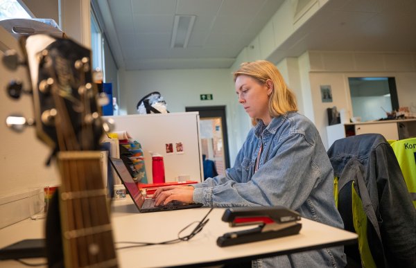 En kvinne med blondt hår og blå olaskjorte sitter på lærerværelset og jobber på en laptop. I forgrunnen, ved siden av pulten står en gitar. I bakgrunnen henger jakker og refleksvester.