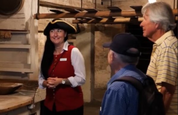 En kvinne med langt, mørkt hår, rød vest, hvit bluse og svart hatt med gul kant står og snakker foran to eldre menn. De befinner seg i et gammelt rom med trebjelker, hyller og rustikt interiør.