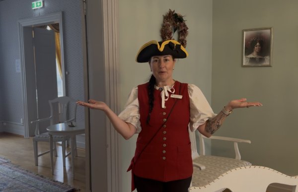 En kvinne med langt, mørkt hår i flette står i et lyst rom med armene ut til sidene og håndflatene vendt oppover. Hun har på seg en rød vest, hvit bluse med puffermer og en svart hatt med gul kant. Rommet er innredet med lyse, gammeldagse møbler, og på veggen henger et innrammet portrett.