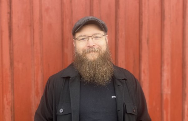 En mann med et langt skjegg. Han har på seg en mørk jakke og t-skjorte, samt en mørk caps og briller. Mannen står foran en rød vegg.